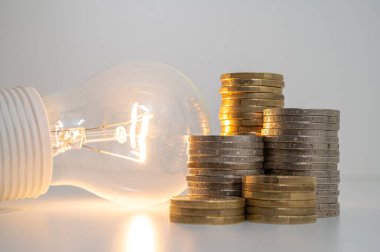 Light bulb turned on, with stacks of coins next to it. Rising electricity tariffs, energy dependency, energy sources and energy supplies.