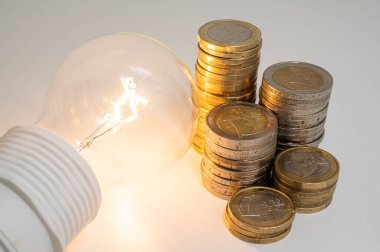 Light bulb turned on, with stacks of coins next to it. Rising electricity tariffs, energy dependency, energy sources and energy supplies.