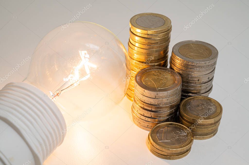 Light bulb turned on, with stacks of coins next to it. Rising