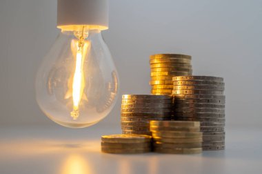 Light bulb turned on, with stacks of coins next to it. Rising electricity tariffs, energy dependency, energy sources and energy supplies.