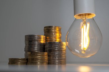 Light bulb turned on, with stacks of coins next to it. Rising electricity tariffs, energy dependency, energy sources and energy supplies.
