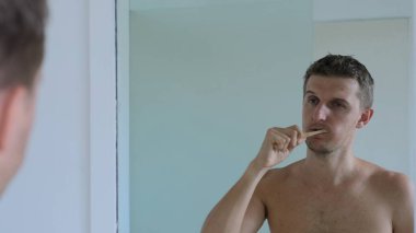 Sleepy man in the morning in the bathroom. A young man stands in front of the mirror naked and cleans the tooth enamel of the teeth. Very careful and attentive to this procedure in the bathroom.