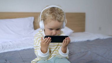A little blond girl is sitting on the bed listening to music in wireless headphones and playing some interesting game on the phone. She is extremely serious and focused.