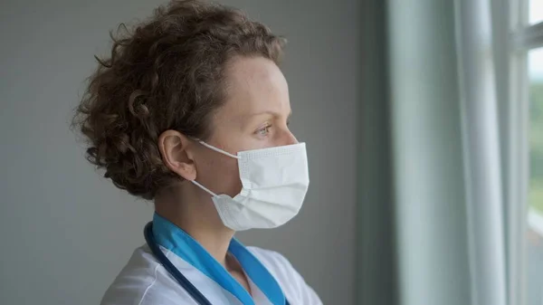 A woman doctor in a mask stands at the window, thinking about the severity of the disease of the patient whom she treats. Treatment of patients infected with a pulmonary infection. Tired nurse.