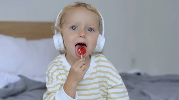 A 3-year-old baby sits on a bed in her mouth with a red sweet candy with the taste of berries. She looks around and sings along to a song singing in her headphones.