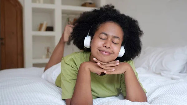 Funny black woman lies on the bed in the room listens to music on headphones and dances. Mixing races results in beautiful people. The routine life of Americans in their free time.