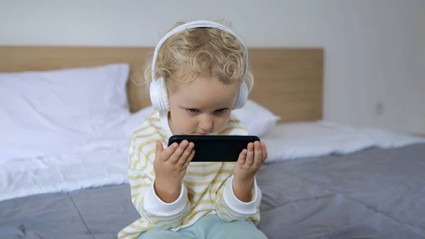 一个金发小女孩正坐在床上 用无线耳机听音乐 并在电话上玩一些有趣的游戏 她是个严肃而专注的人 图库图片