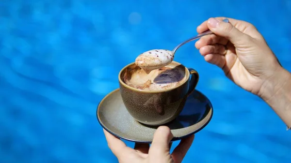 在蓝水的背景下 女性的手捧着一杯泡沫咖啡 慢动作的勺子搅动着卡布奇诺泡沫 香浓的咖啡能让人充满活力 图库照片