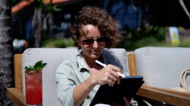 Freelance work from anywhere in the world via the Internet. Curly girl in sunglasses works as an illustrator sits on the beach in tropics with a soft drink is engaged in work using a graphics tablet