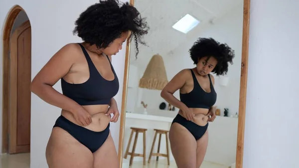 An African-American woman in black underwear in front of a mirror draws in a fat belly, and cannot believe that her body has become so fat. Obese women from eating fast food around the world.