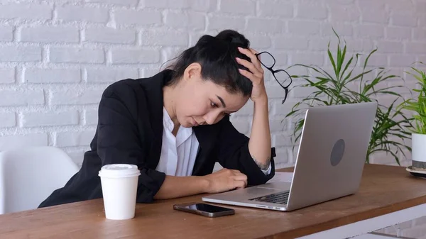 Tired woman works at the office with a laptop. The woman has a headache from overwork, she takes off her glasses and sighs heavily. On the table is a laptop, smartphone and takeaway coffee