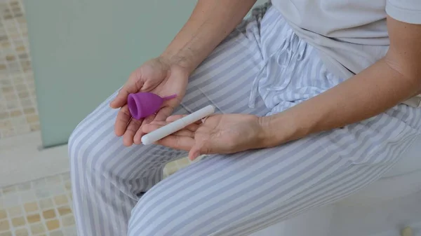 In the restroom for natural needs sits a woman on a toilet. The woman is dressed in home clothes, two personal care objects lie on her palms, a choice between a tampon and a menstrual cup.