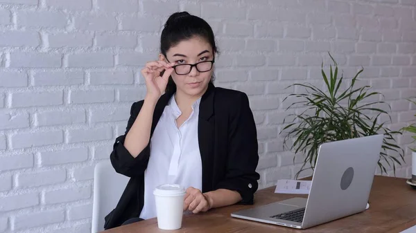 An employee of the office, dressed in stylish clothes with a strict appearance, her outfit inspires confidence and respect, was distracted from work, lowering her glasses and looking closely.
