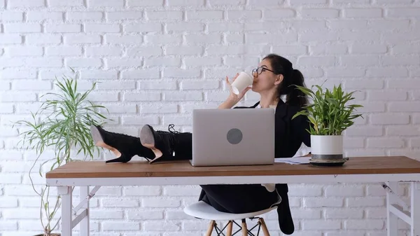 The office employee looks immaculate. The design of the office is made in a modern style. Decor flowers. The employee herself decided to rest, putting her feet on the table and drinking a drink.