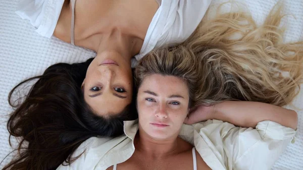 Top view of women who are lying on the bed looking at the camera. A couple of beautiful lesbians who spend time together. Friends who love each other. Blonde and brunette are made for same-sex love.