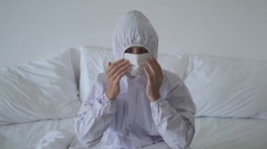 Beyaz takım elbiseli ve koronavirüse karşı maske takan bir kadın yatakta yatıyor. Koronovirüs paniği ve salgın sırasında hastalığa karşı aşırı koruma. Enfeksiyon kapma korkusu.