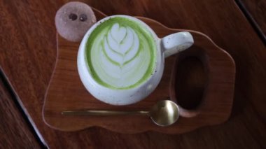 Top down view of a beautifully served cup of matte green tea with an ingenious pattern on the milk froth. The cup stands on a wooden tray in the same color scheme as the table.