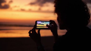 Silhouette of a womans head with shoulders against the backdrop of a twilight sunset. A woman holds a smartphone in her hands and takes pictures of the sun hiding behind the horizon.