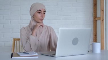 A beautiful Muslim woman in a hijab sits at a table, uses a laptop, types on a keyboard. Freedom in the Islamic world in relation to women. Modern History of the Islamic World. High quality 4k footage