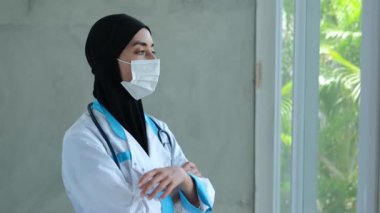 A Muslim woman doctor stands by a window in the office. Removes the medical mask from the face. He looks out the window. Touches his forehead. The Muslim woman doctor has a tired look.