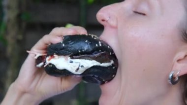 Close-up of a woman takes a big bite from an appetizing black burger with sauce and chews delicious food with pleasure. An unhealthy diet leads to diseases of the stomach and heart.