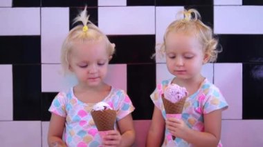 Two adorable twin girls eat ice cream together. The pleasure of children from eating sweets. Two babies of three years old eat ice cream in waffle cones. Happy emotions of children.