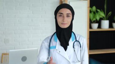 A female doctor in a medical gown and a black hijab shows a thumbs up, a Muslim woman at her workplace in an office in a hospital, a Muslim woman works as a nurse, helps people. 