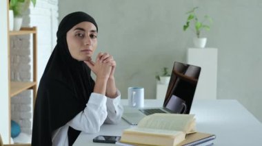 A beautiful Muslim woman dressed in light clothes and a black hijab sits at a table. Online university education On the table is a laptop, there is an open book, a phone and other items.