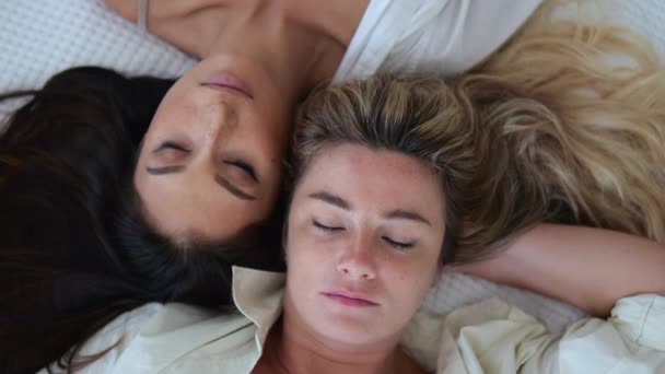 ベッドの中で二人の美しい女性のトップビューは 彼らの頭がお互いにもたれて眠る 彼らの目を閉じて ベッドの中でお互いを感じるブロンドとブルネットのモデルの肖像画 — ストック動画