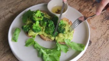 A healthy breakfast of toast with avocado and broccali cabbage sauce. A white plate of fried toast with avocado spread and green broccoli cut with a knife, dressing sauce and lettuce.