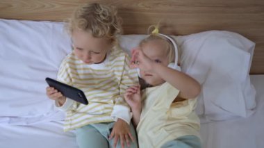 Blonde blue-eyed sisters are lying in their parents bed, watching an interesting cartoon together on the phone. They learned to use gadgets at an early age.