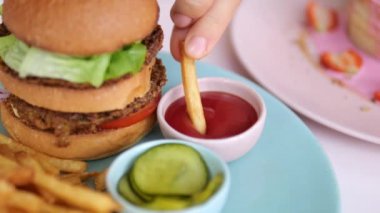 Close-up of a plate with a delicious hamburger and garnish in the form of french fries or chips. Take one piece of french fries with your fingers and dip in tomato ketchup sauce. Junk food.