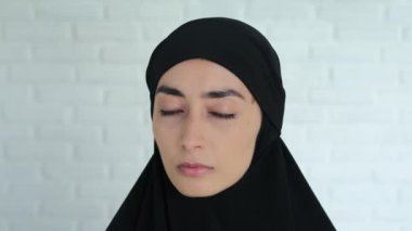 Siyah tesettürlü genç bir kadının beyaz tuğladan bir duvara baktığı ve kayıtsız, ağır bir bakışla kameraya baktığı yakın plan görüntüsü. Müslüman kadınların ihlali.