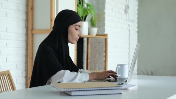 美しいイスラム教徒の女性学生がノートパソコンのキーボードを入力しています 黒いヒジャーブの女性が大学の記事を入力しています イスラームにおけるジャーナリスト女性としての仕事 — ストック動画