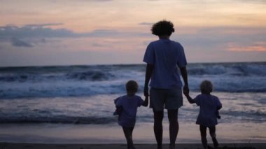 In the rays of the setting sun, a single mother leads two children to the water holding their hands. The ocean with large waves runs to the sandy shore of the beach where a family walks.
