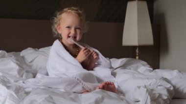 A sweet little girl of two years old sitting on a bed just woke up. The girl brushes her teeth from an early age, parents accustom the child to oral hygiene. Morning procedures of the baby.