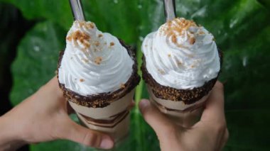 Two appetizing milkshakes with whipped cream with metal tubes for a cocktail are held in hands against the background of a green leaf of a tropical palm tree. Drinks on holiday at the hotel.