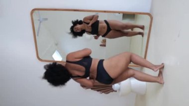 A vertical video of an African American plus size model dancing at home in front of a mirror in black lingerie. A woman does not pay attention to excess weight, she loves herself for who she is.