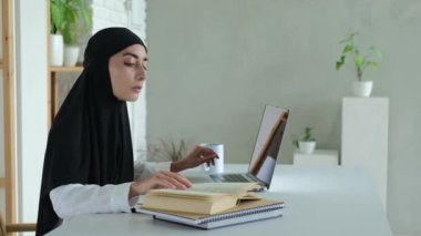 A Muslim female teacher reads a book and types learning material on a laptop keyboard. The rights of Muslim women to education. A woman in a black hijab conducts remote training.