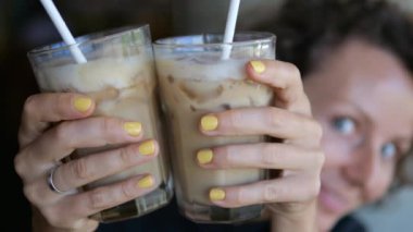 In the foreground, close-up, a woman holds two glasses with a chocolate cocktail with milk and ice in her hands. She peeps out laughing, now from one side, then from the other.