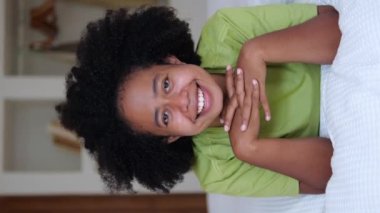 A cheerful black woman with an afro hairstyle lies on the bed and smiles with a snow-white smile and sings songs. Portrait of an Afro Asian woman in a home interior. vertical video.