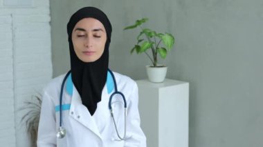 An Arab woman stands in the office dressed in a black hijab, a medical gown with blue inserts and a medical device hung over the hijab. Calm and self-confident female doctor in Islam.