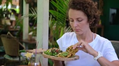 A woman is sitting at a table in a cafe. She holds a small pineapple-shaped tray in her hand. She takes an eco-friendly waffle from the tray with her other hand. She is hungry and eats greedily.