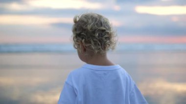 Shot from behind. A small blond child with an angelic appearance walks along the seashore against the backdrop of an evening sunset. She slowly turns around and looks at the one who called out to her.