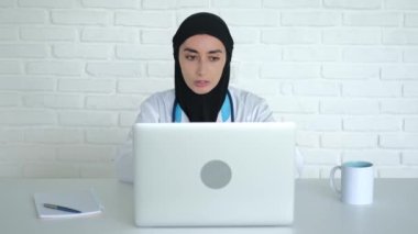 Online consultation of the patient, in a Muslim clinic. A doctor in a hijab and a medical uniform sits at a table in a hospital, looks into a laptop camera, communicates with a patient via video link.