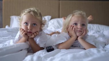 Shooting sisters from the foreground close-up. They lie on the bed and watch TV which is above their eye level. The screen shows childrens content. The TV remote is next to them.