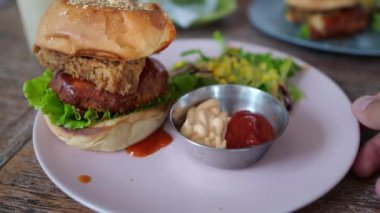 A medium-sized hamburger lies on a white plate. The top bun is sprinkled with sesame seeds. He looks appetizing. Nearby is a portion of salad and two sauces.