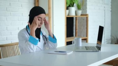 A Muslim woman, a doctor, sits on a chair. A tired doctor with a headache at his workplace helps the sick by herself. On the desktop there is a laptop and a notebook with a pen for notes.