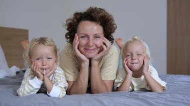 Two cute blond girls have fun together with their mother. They lie on a huge bed. The little ones hold lollipops in their mouths, smiling cheerfully. So carefree they spend their holidays.