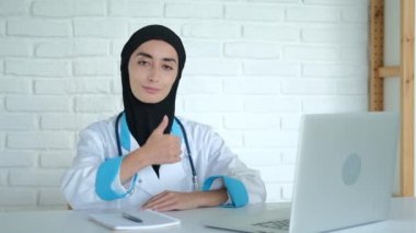 A happy Muslim woman can work at her favorite job in the clinic. A woman in a hijab sits at a table in a hospital showing thumbs up, rejoices that she can help people.